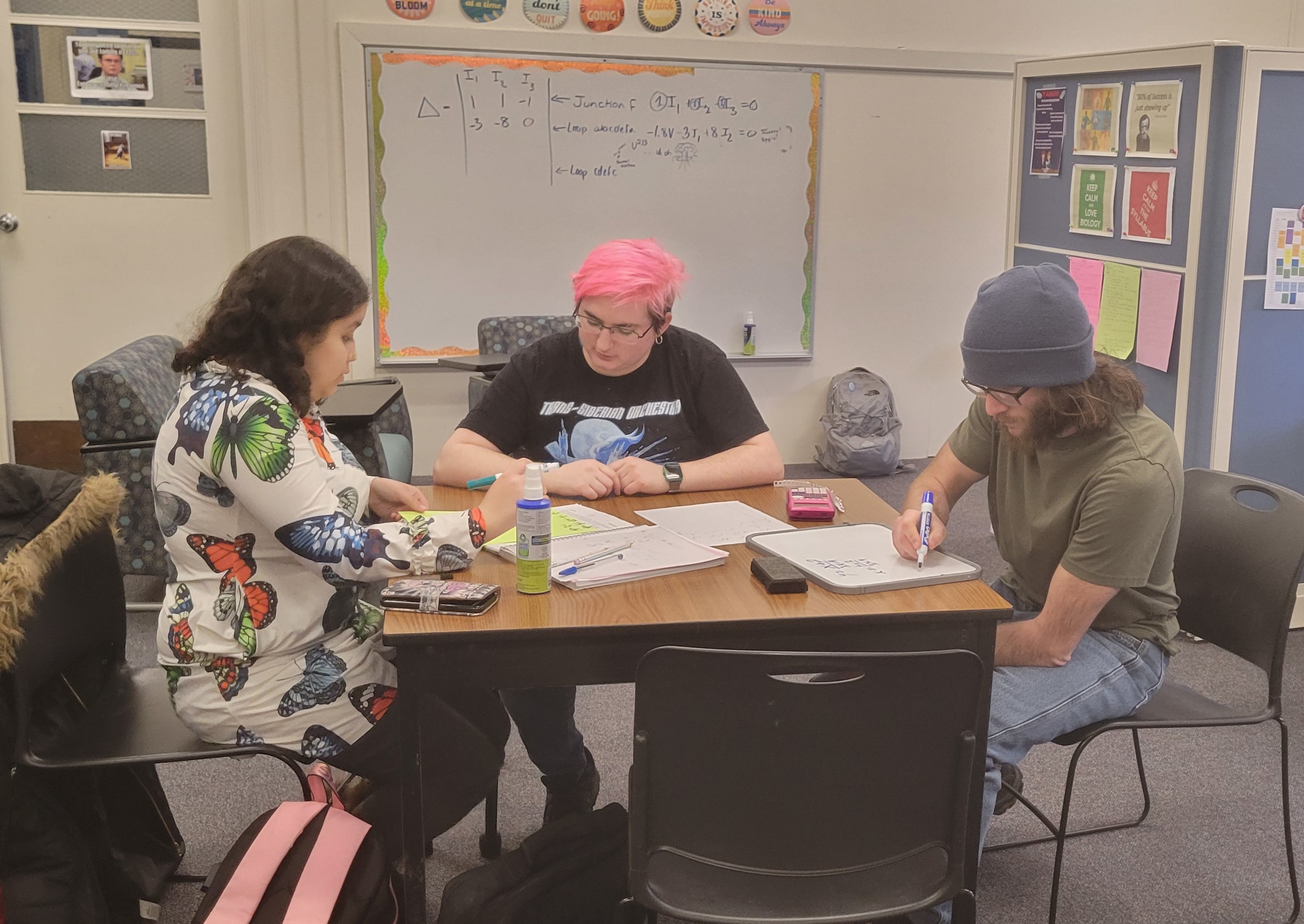 Three students sitting at a square table writing on dry erase boards doing school work